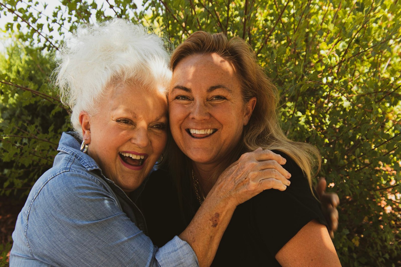 An adult daughter and her smiling older mother embracing outdoors in warm afternoon light