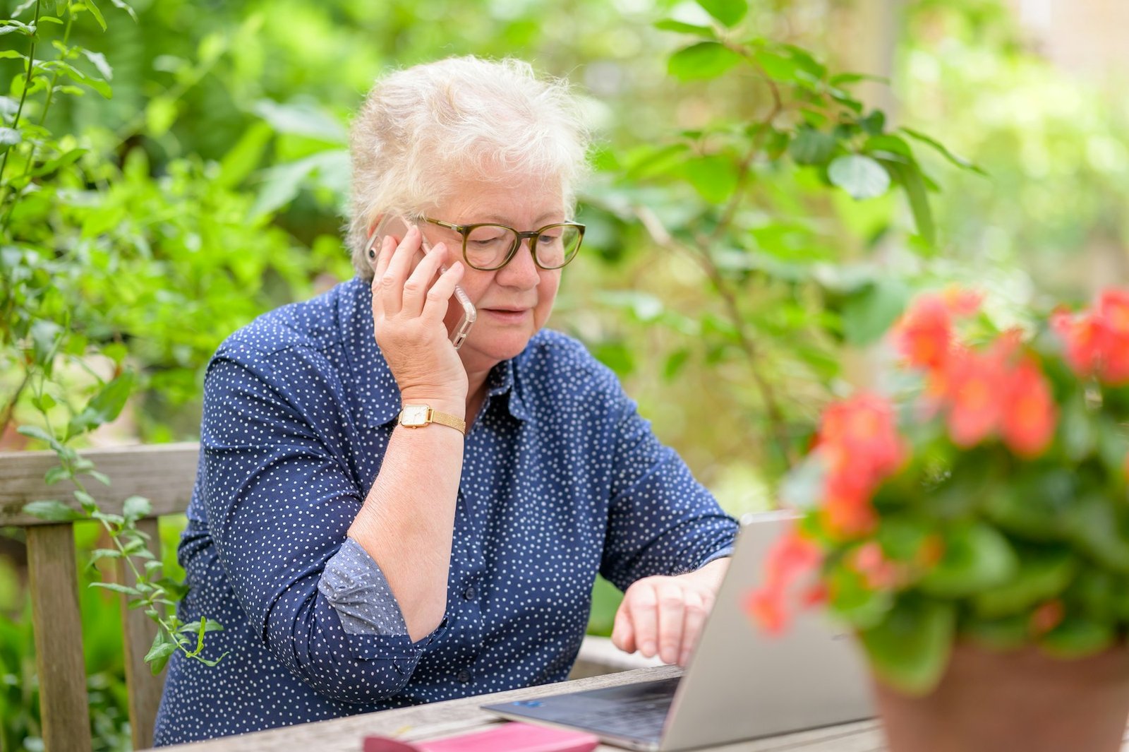 An older woman with glasses researching on a laptop beside flowering plants